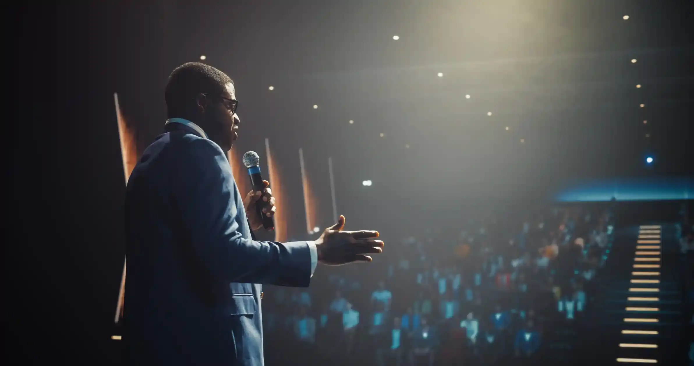Home Page 1 A speaker in a suit holding a microphone on a stage, facing a large audience in a dimly lit conference auditorium, suggesting a keynote address or professional event.