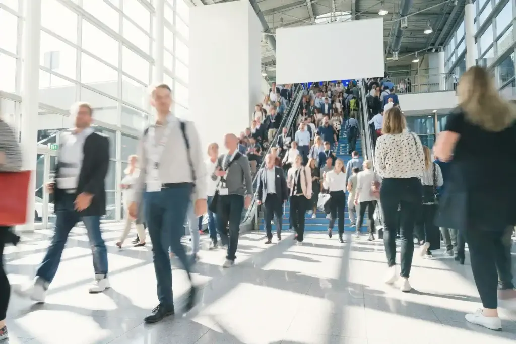 Home Page 5 Crowd of business professionals walking through a large convention center near escalators at a trade show or corporate event, with bright natural light from tall windows.